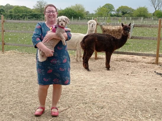 Emma Lowery stands in an Alpaca pen, with a white and brown alpaca standing behind her, by their hay rack, looking at the camera, and she is standing in the foreground holding a small pale brown dog in her arms. 